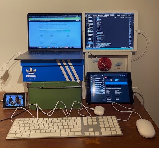 Banco escolar em duas cadeiras. Em cima da mesa da escola, há caixas de sapatos apoiando um laptop e dois iPads ao redor.